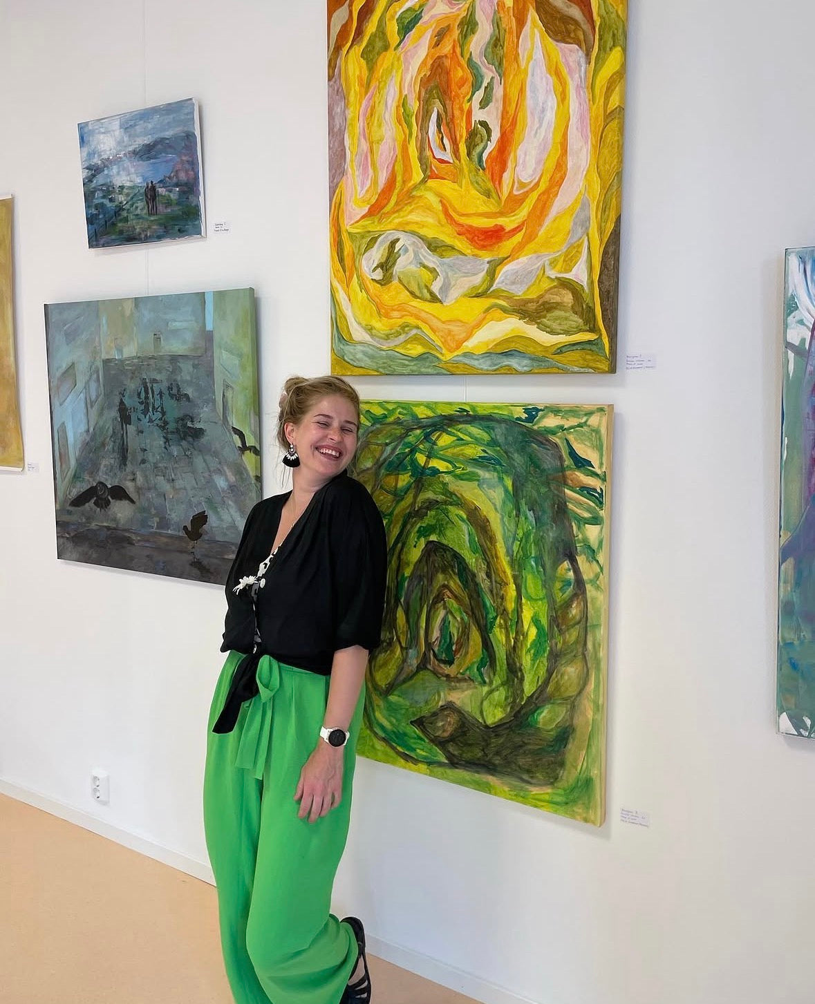 Foto av kunstneren foran to av kunstverkene på utstilling i Galleri Minneparken 2022. Du ser to bilder som henger over hverandre, det nederste grønt det øverste gult. 